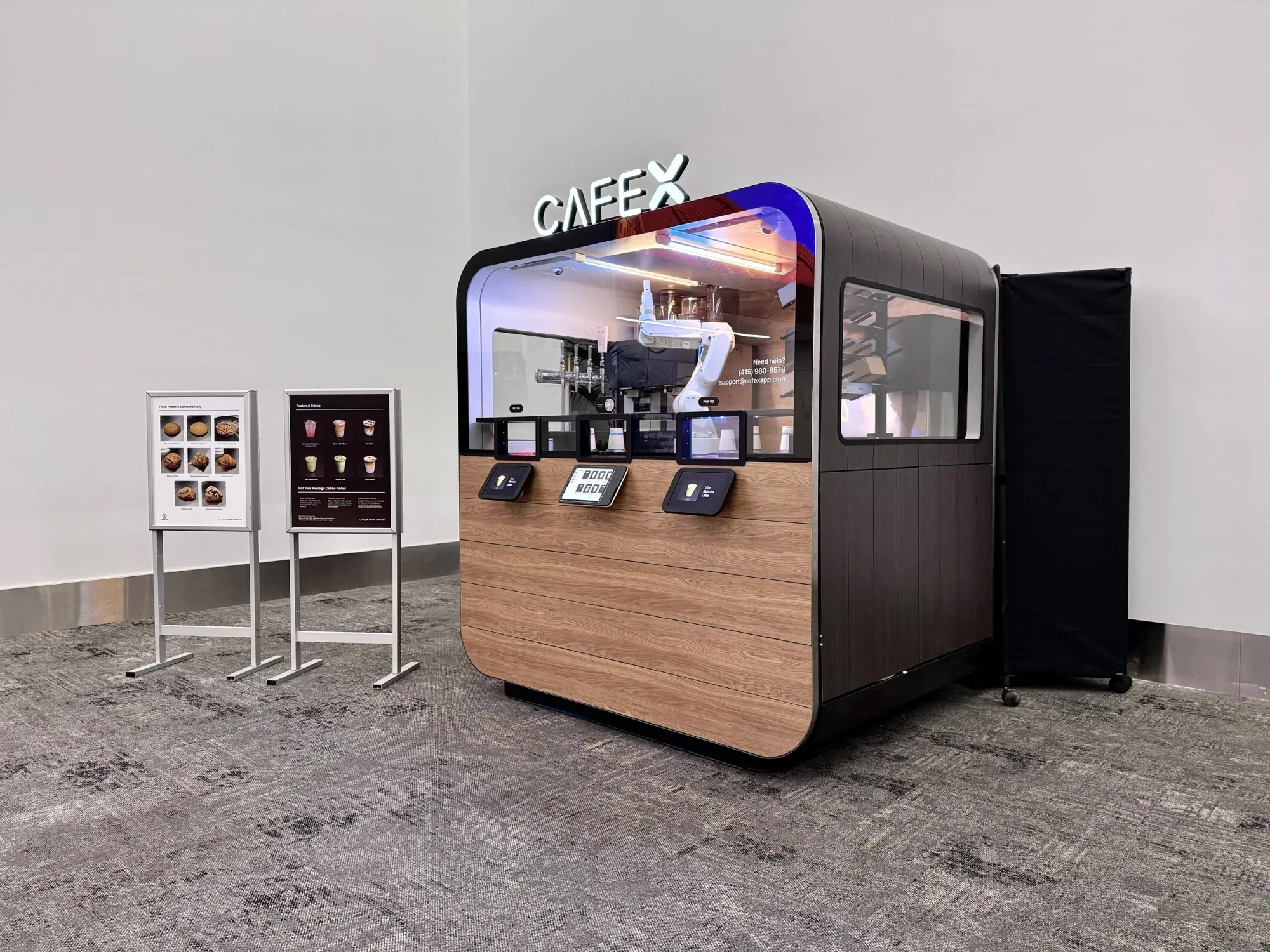 A wood-paneled, fully enclosed kiosk in a carpeted area of a building. There are small slots in the glass for customers to retrieve their drinks, and tablets presumably for ordering. Behind the glass is a large robotic arm, like what you might expect to see on an assembly line.