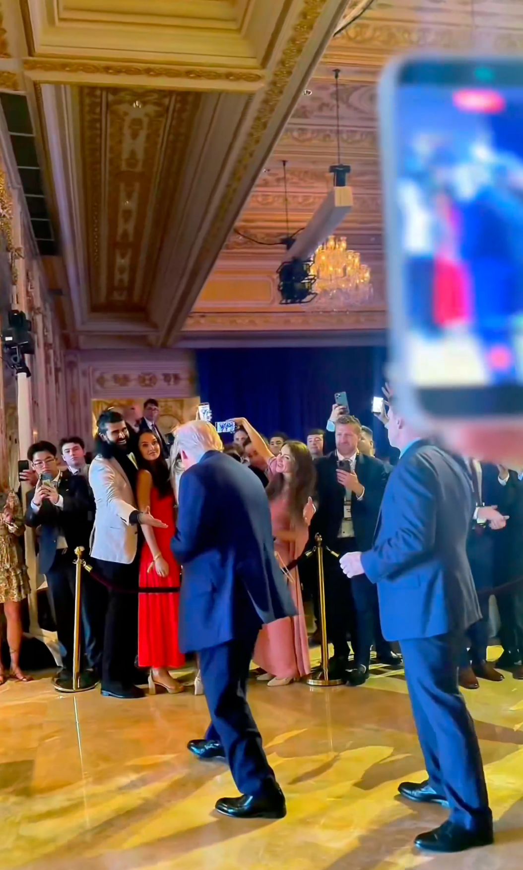 Trump steps towards a man reaching out to shake his hand from behind a cordon. A crowd of other attendees are mostly taking photos with their cell phones.