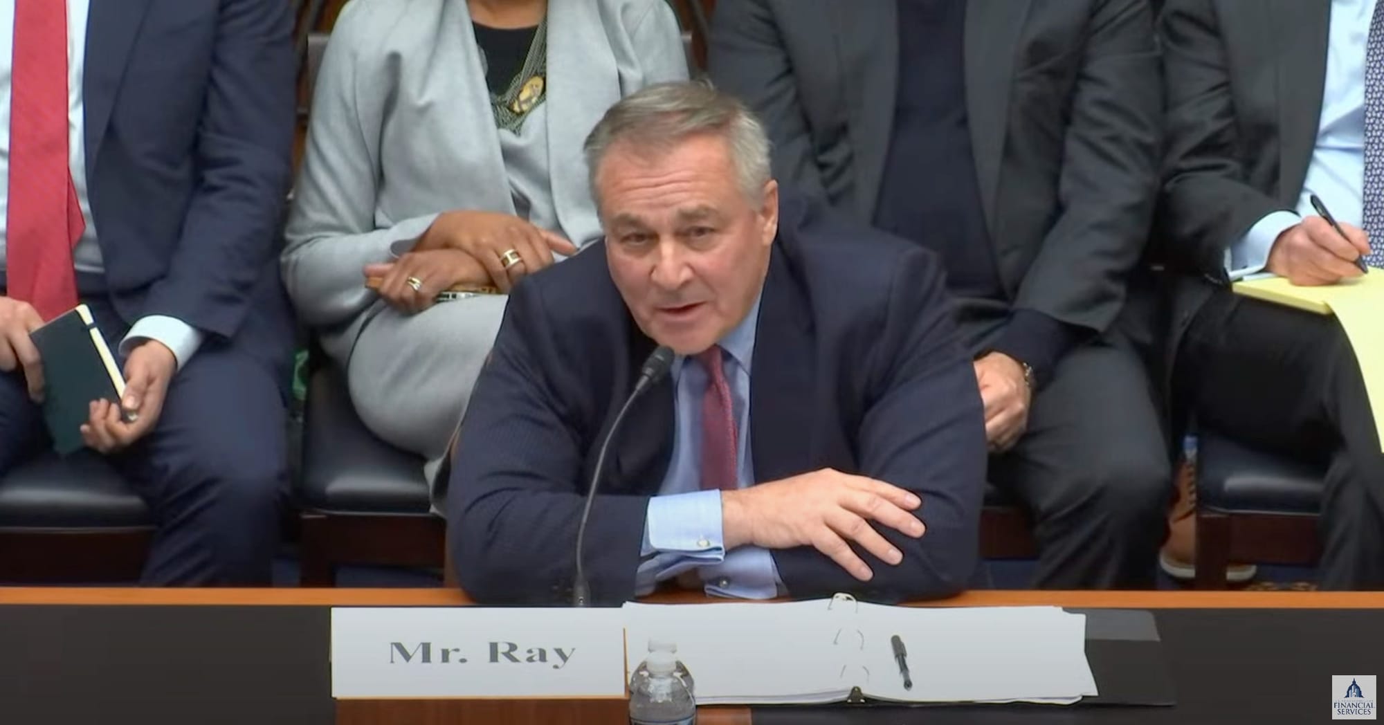 John Jay Ray III sits with his arms folded on the table in front of him, speaking into a microphone at the House FSC hearing.