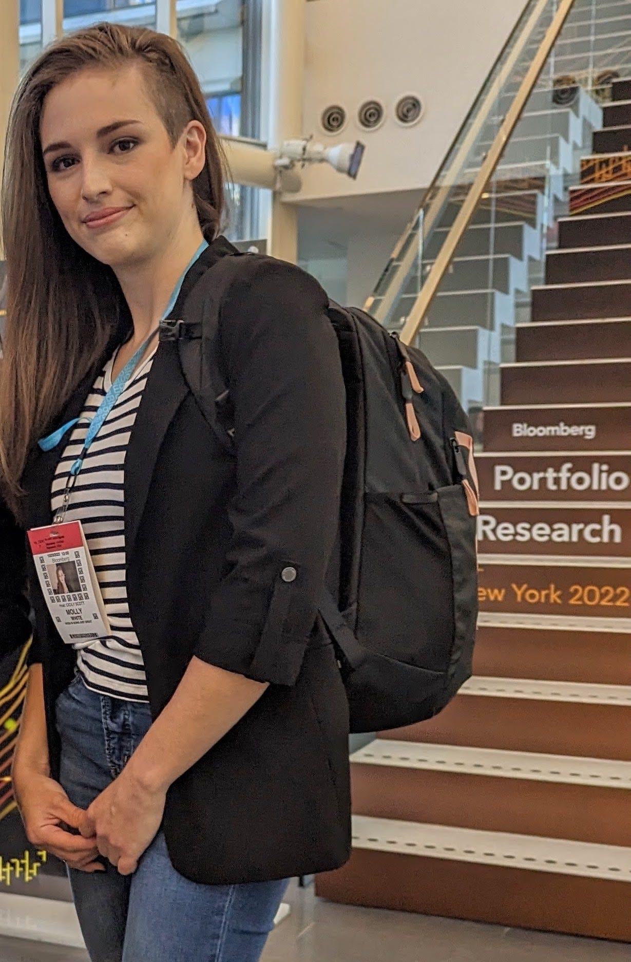 Photo of Molly standing in an office building. She's wearing a white and navy striped shirt, a black blazer, and jeans. Behind her is a large staircase with the text "Bloomberg Portfolio Research New York 2022" printed on the risers.