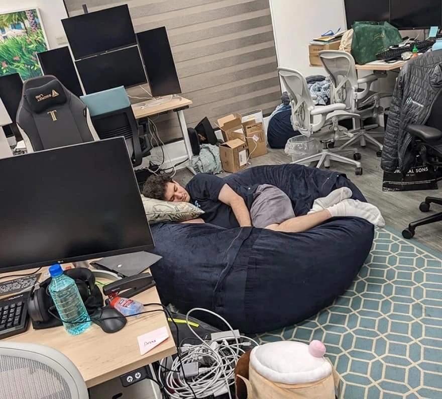 Sam Bankman-Fried sleeps curled up on a large navy blue beanbag in a cluttered office, surrounded by computer monitors. He's wearing a t-shirt, shorts, and oversized white gym socks.