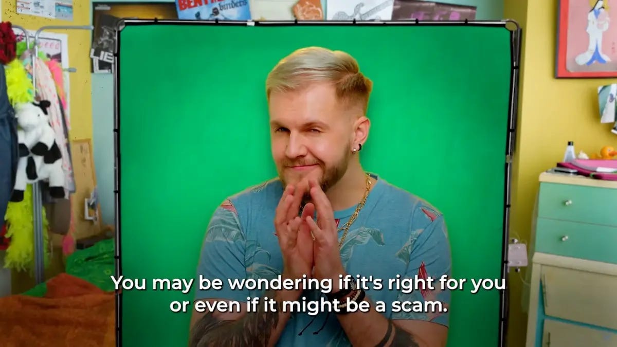 A man with blond hair and a short beard makes a devious squinting face with his hands steepled in front of him. He's sitting in front of a green screen in a bedroom, and wearing a gold necklace and blue patterned t-shirt.