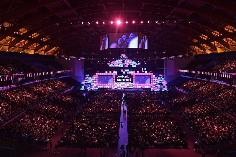 An image of a full Altice Arena. The stage has pink and blue light blocks forming a backdrop, with the Web Summit logo in the middle. There is a jumbotron displaying faces of some audience members.