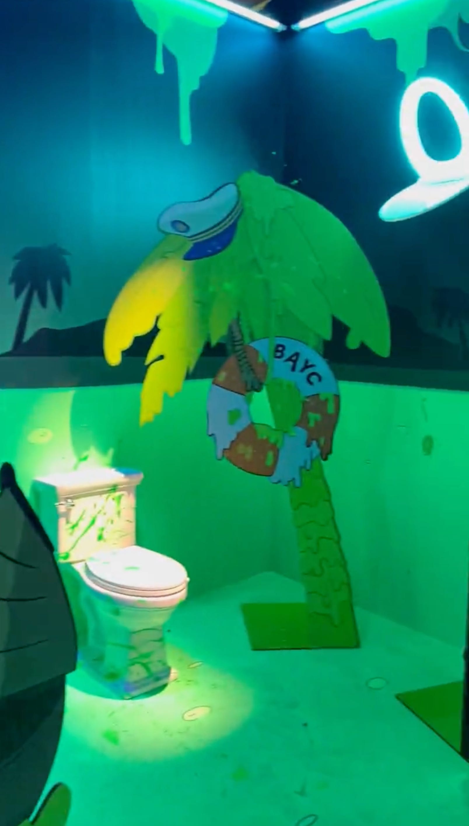 A room with a toilet, a cardboard cutout of a palm tree with a sailor hat and BAYC life ring. The walls are adorned with toilet seats and the room is lit with blacklights.
