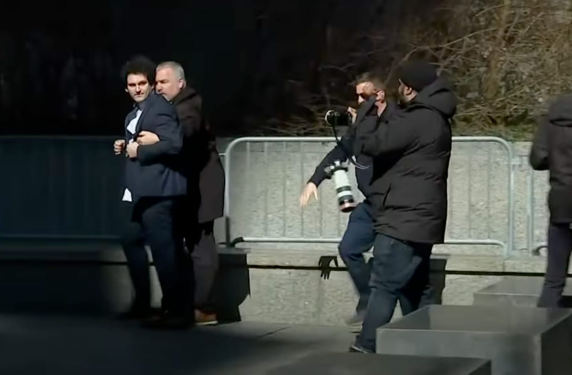A bodyguard walks next to Sam Bankman-Fried with his arms underneath SBF's arms. They have just exited a crowd of photographers. SBF is wearing a navy suit and is looking back at the photographers while walking forwards.