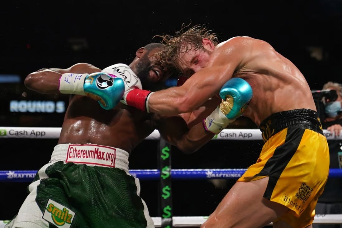 Boxer Floyd Mayweather hits Logan Paul in the chest with an uppercut. Mayweather is wearing shorts with a large EthereumMax logo across the front waistband.