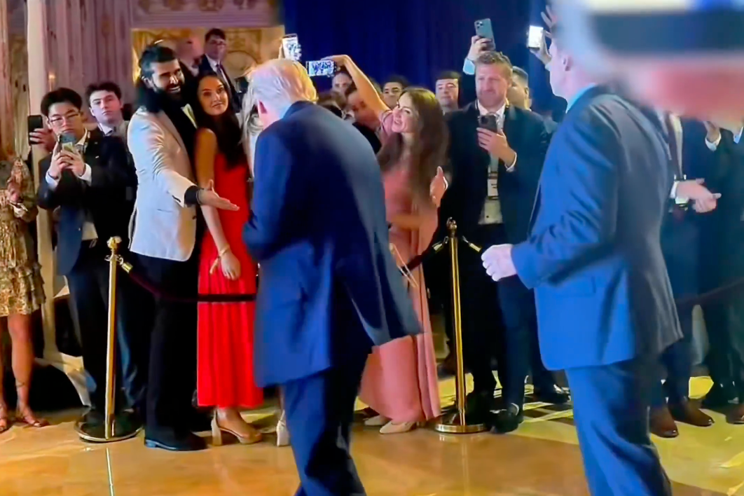 Trump, photographed from behind, steps towards a man reaching out to shake his hand from behind a cordon. A crowd of other attendees are mostly taking photos with their cell phones.