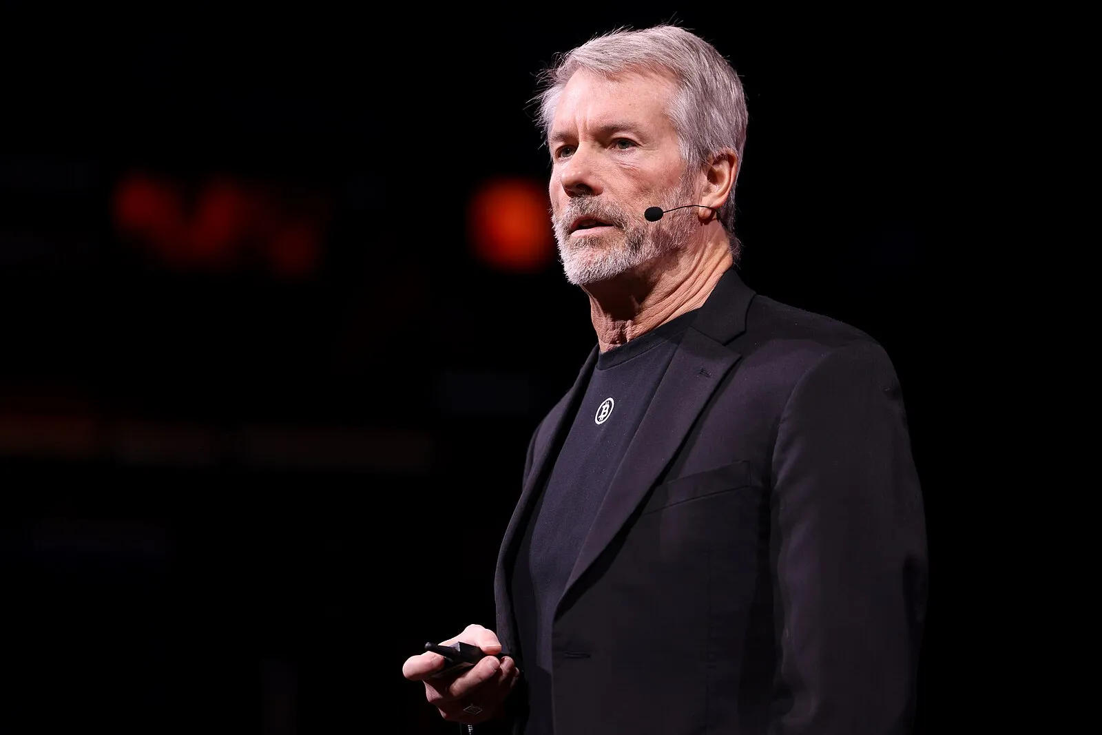Michael Saylor standing on-stage. He’s wearing a black suit jacket over a t-shirt with a white bitcoin logo on it