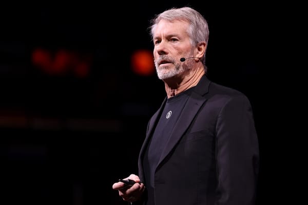 Michael Saylor standing on-stage. He’s wearing a black suit jacket over a t-shirt with a white bitcoin logo on it