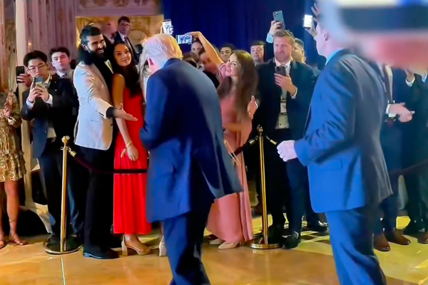 Trump, photographed from behind, steps towards a man reaching out to shake his hand from behind a cordon. A crowd of other attendees are mostly taking photos with their cell phones.