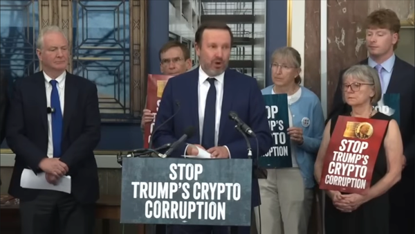 Senator Chris Murphy (D-CT) speaks at a podium labeled with “Stop Trump’s Crypto Corruption”. Behind him stands Senator Van Hollen (D-MD) and a group of other people.