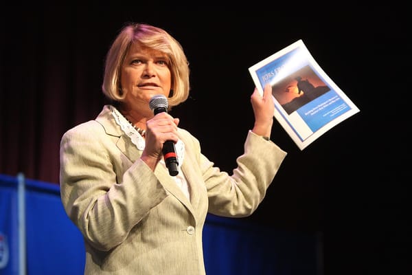 Senator Cynthia Lummis speaks into a microphone on a stage, holding up a paper