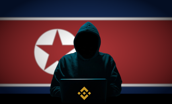 A person in a black hoodie with their face in shadow sits at a computer with the Binance logo on the back. Behind them is a DPRK flag.